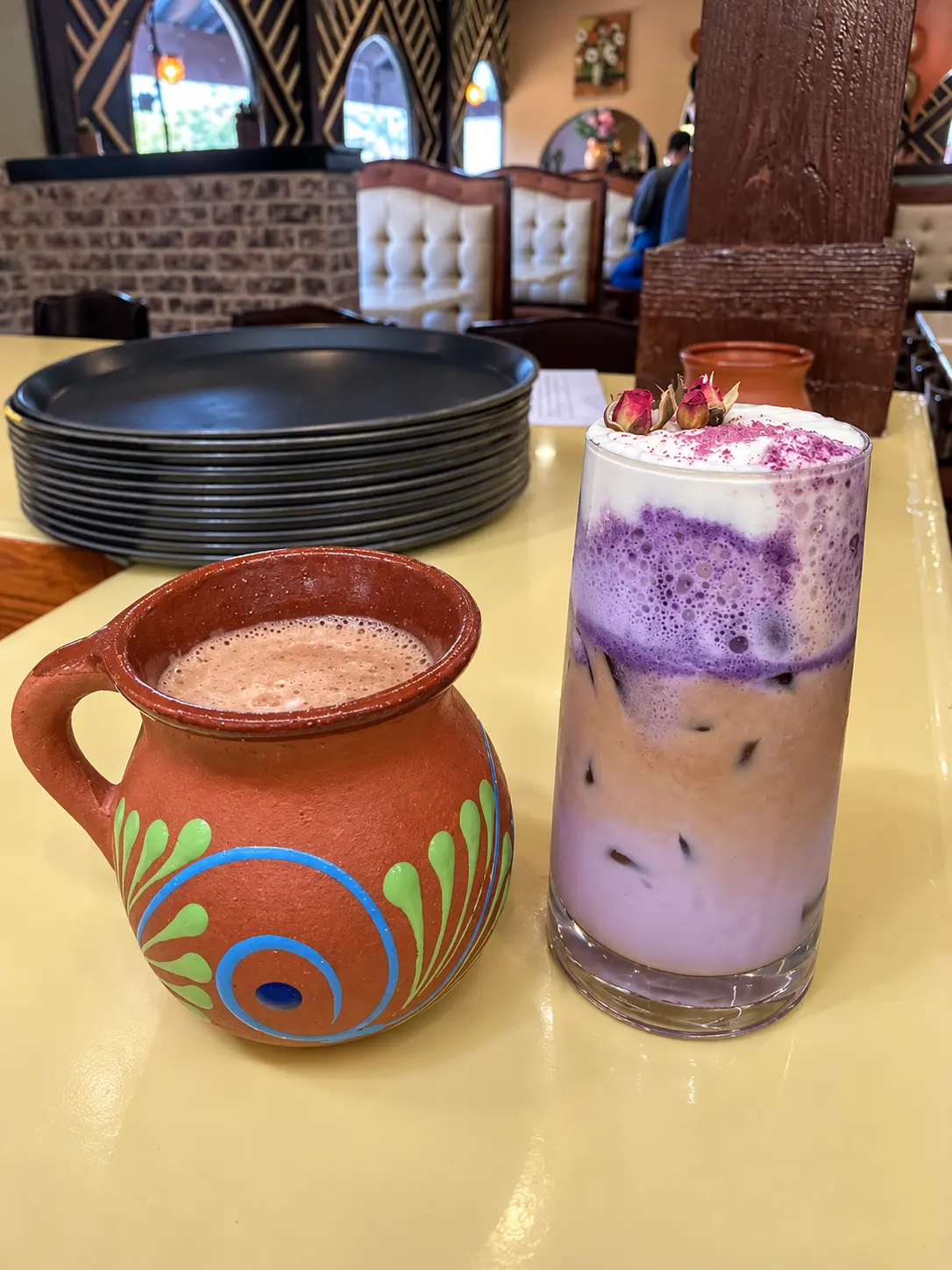 A clay mug of coffee beside a purple iced latte topped with rosebuds